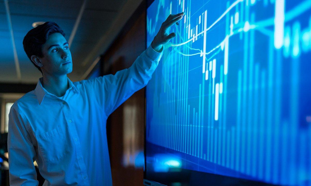 Male college student touching large computer screen graphs.