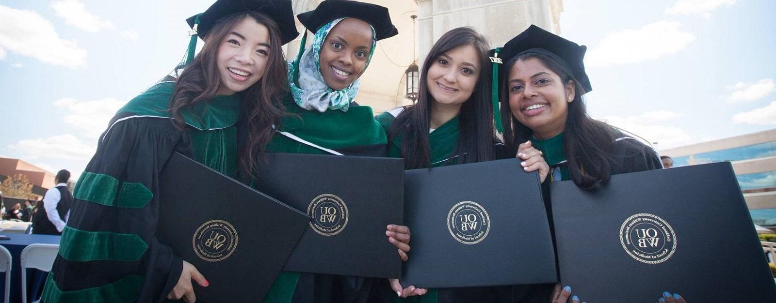 Four new OUWB graduates stand before the Elliot Tower wearing caps and gowns and holding their diplomas.